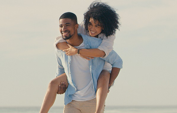 Un couple sur la plage qui regarde au loin, l'homme portant la femme sur son dos.